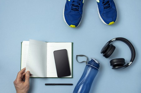 Male hands, headphones,  sneakers, bottle of waters, smartphone, notebook diary with marker on blue background.の写真素材