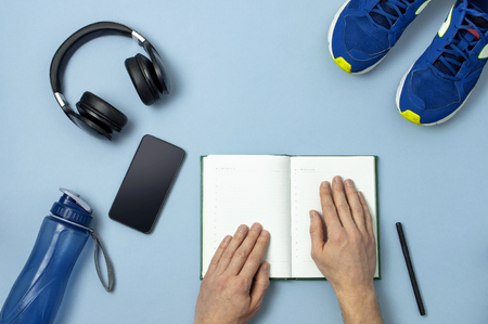 Male hands, headphones, blue sneakers, bottle of waters, smartphone, notebook diary with marker on blue background Top view flat lay copy space. Concept of active lifestyle sports fitness health.の写真素材