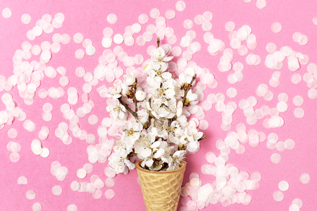 Waffle cone with bouquet of spring white flowers and round confetti on pastel pink background. Festive holiday background, concept of party, birthday. Creative spring minimal style Flat lay top view.の写真素材