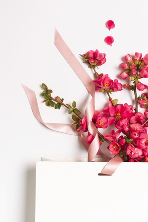 Flat lay White gift bag package with ribbon and spring pink flowers on light background top view. Greeting card with delicate flowers Pink floral background. Spring minimal concept. Beauty fashion.の写真素材