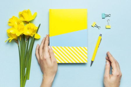 Women's hands, open blank notebook, pen, clips, spring flowers daffodils narcissus on blue background. Female desktop, Office desk, spring concept. Flat lay, top view, copy space. Template for blog.の写真素材