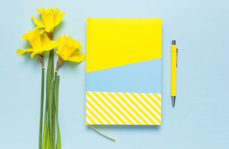 Yellow-blue notebook, pen, clips, spring flowers daffodils narcissus on blue background. Female desktop, Office desk, spring concept. Flat lay, top view, copy space. Template for feminine blog.の写真素材