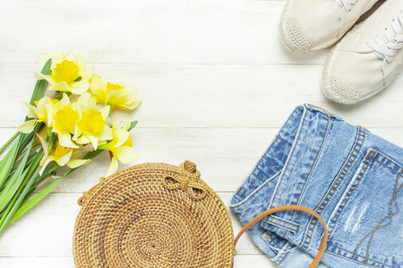 Fashionable natural organic round rattan bag, denim shorts, beige women's espadrilles, yellow narcissus daffodil flowers on light wooden background flat lay. Summer fashion concept. Ecobags from Bali.の写真素材