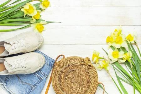 Fashionable natural organic round rattan bag, denim shorts, beige women's espadrilles, yellow narcissus daffodil flowers on light wooden background flat lay. Summer fashion concept. Ecobags from Bali.の写真素材