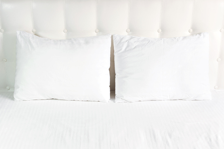 Soft white quilted pillow in bed on the background of white leather quilted headboard. Clean pillow, part of bed close-up, comfort. Quilted headboard background, bedding mockup design template.の写真素材