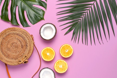 Summer fashion flat lay. Round trendy rattan bag, tropical palm leaves, monster leaf, coconut, orange on pink background. Top view, copy space. Trendy bamboo bag. Creative background, vacation concept.の写真素材