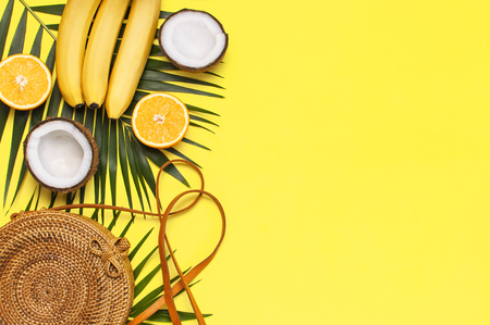 Summer fashion flat lay. Round trendy rattan bag, tropical palm leaves, coconut, orange, bananas on yellow background. Top view, copy space. Trendy bamboo bag. Creative background, vacation concept.の写真素材