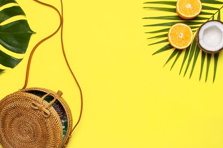 Summer fashion flat lay. Round trendy rattan bag, tropical palm leaves, monster leaf, coconut, orange on yellow background. Top view copy space. Trendy bamboo bag. Creative background vacation concept.の写真素材