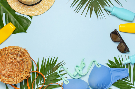 Summer composition flat lay. Frame of Round trendy rattan bag straw hat sunglasses tropical palm leaves swimsuit sunscreen seashells on blue background. Top view copy space. Creative fashion backdrop.の写真素材