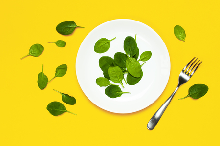 Fresh green spinach leaves on a white plate fork on bright yellow background flat lay top view copy space. Baby young spinach leaves, healthy food, diet. Creative food conceptの写真素材