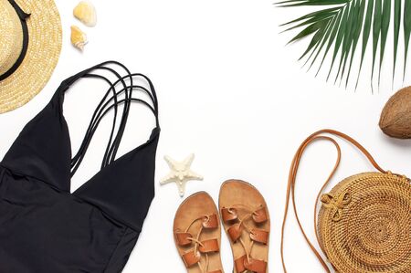 Woman's beach accessories flat lay. Round trendy rattan bag straw hat black swimsuit leather sandals tropical palm leaves coconut seashells on white background. Top view copy space. Summer backdrop.の写真素材