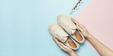 Female hands hold beige fashionable espadrilles with spring flowering branch on pastel blue pink background. Creative concept of spring shoes, fashion blog or magazine concept Flat lay top view.の写真素材