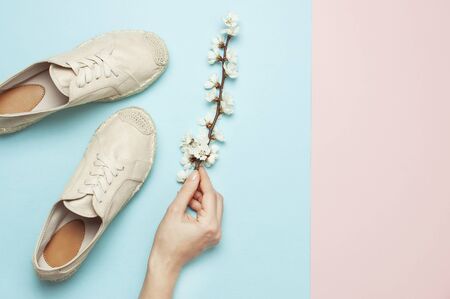 Female hands hold beige fashionable espadrilles with spring flowering branch on pastel blue pink background. Creative concept of spring shoes, fashion blog or magazine concept Flat lay top view.の写真素材