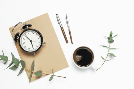 Flat lay cup of black coffee, black alarm clock, notebook, diary, eucalyptus on white background top view copy space. Minimalistic food concept, Feminine desk workspace, time to work coffee background.の写真素材