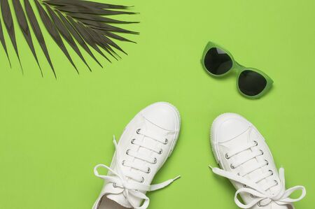 White female fashion sneakers, trendy sunglasses, tropical leaves on green background. Flat lay top view copy space. Women's shoes, accessories. Stylish white sneakers. Creative summer concept.の写真素材