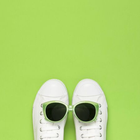 White female fashion sneakers and trendy sunglasses on green background. Flat lay top view copy space. Women's shoes, accessories. Stylish white sneakers. Creative concept Fashion blog or magazine.の写真素材