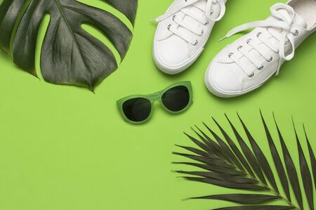White female fashion sneakers, trendy sunglasses, tropical leaves on green background. Flat lay top view copy space. Women's shoes, accessories. Stylish white sneakers. Creative summer concept.の写真素材