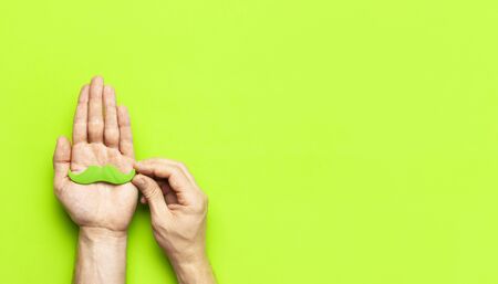 Creative party decoration concept Men's hands hold green mustache, props for photo booths, carnival parties on green background top view flat lay copy space Father's day Men's health awareness month.の写真素材