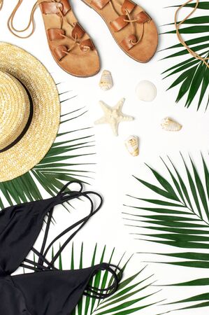 Woman's beach accessories flat lay. Round trendy rattan bag straw hat black swimsuit leather sandals tropical palm leaves seashells on white background. Top view copy space. Summer backdrop.の写真素材