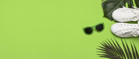 White female fashion sneakers, trendy sunglasses, tropical leaves on green background. Flat lay top view copy space. Women's shoes, accessories. Stylish white sneakers. Creative summer concept.の写真素材