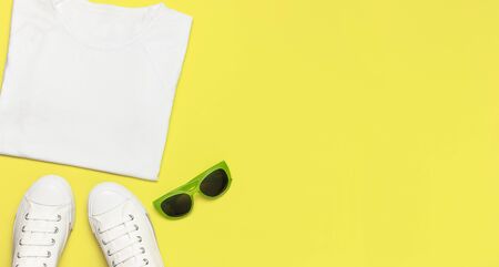White female fashion sneakers, trendy sunglasses, white T-shirt on yellow background. Flat lay top view copy space. Women's shoes, accessories. Stylish white sneakers. Creative concept Fashion blog.の写真素材