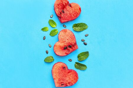 Creative summer food concept. Juicy slices of ripe red watermelon in the shape of a heart and mint leaves on blue background. Flat lay, top view, copy space. Watermelon pattern. Love for watermelon.の写真素材