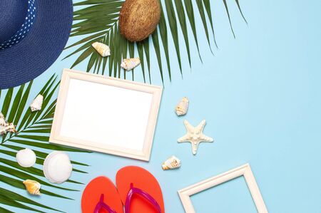 Summer fashion flat lay. Blue hat coral flip flops tropical palm leaves sunscreen white photo frame coconut seashells starfish on blue background. Top view copy space. Creative vacation background.の写真素材