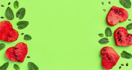 Creative summer food concept. Frame of Juicy slices of ripe red watermelon in the shape of a heart and mint leaves on green background. Flat lay, top view, copy space. Watermelon pattern.の写真素材