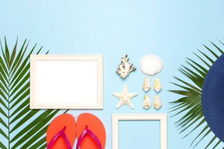 Summer fashion flat lay. Blue hat coral flip flops tropical palm leaves sunscreen white photo frame coconut seashells starfish on blue background. Top view copy space. Creative vacation background.の写真素材