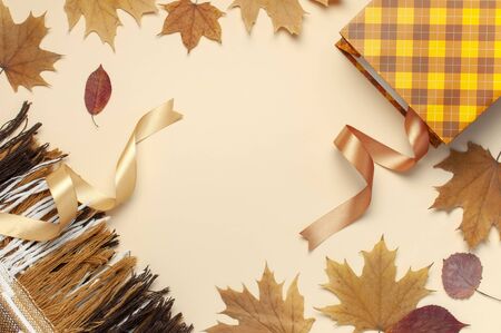 Autumn, fall concept. Checked orange gift paper bag, warm cozy blanket plaid, autumn dried leaves, holiday ribbons on pastel beige background.の写真素材