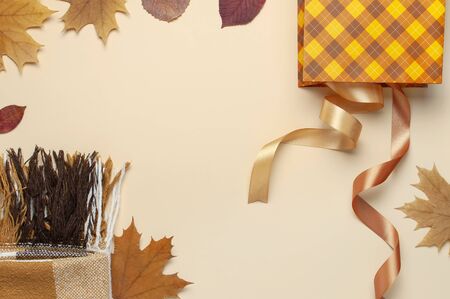 Autumn, fall concept. Checked orange gift paper bag, warm cozy blanket plaid, autumn dried leaves, holiday ribbons on pastel beige background.の写真素材
