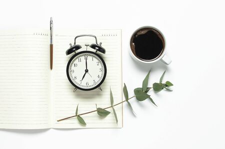 Flat lay cup of black coffee, black alarm clock, notebook, diary, eucalyptus on white background top view copy space.の写真素材
