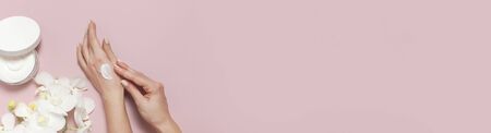Young woman moisturizes her hand with cosmetic cream lotion opened container with cream body milk White Phalaenopsis orchid flowers on pink background Flat lay top view minimalism style Beauty concept.の写真素材