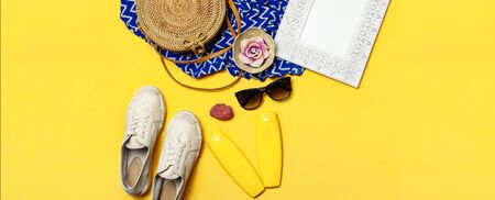 Female summer fashion background. Clothes and accessories on yellow background. Blue dress, round rattan bag, espadrilles shoes, sunglasses, sunscreen, wooden picture frame. Flat lay top view.の写真素材