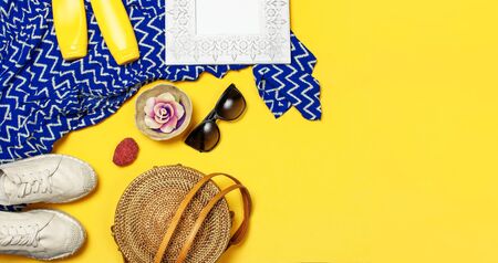 Female summer fashion background. Clothes and accessories on yellow background. Blue dress, round rattan bag, espadrilles shoes, sunglasses, sunscreen, wooden picture frame. Flat lay top view.の写真素材
