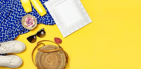 Female summer fashion background. Clothes and accessories on yellow background. Blue dress, round rattan bag, espadrilles shoes, sunglasses, sunscreen, wooden picture frame. Flat lay top view.の写真素材