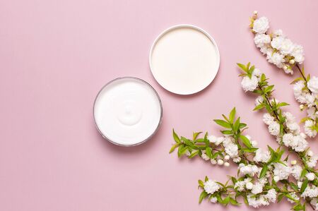 Beauty Spa concept. Opened plastic container with cream and spring White flowers on pink background Flat lay top view. Herbal dermatology cosmetic hygienic cream, organic cosmetic Natural.の写真素材