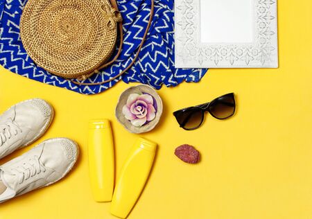 Female summer fashion background. Clothes and accessories on yellow background. Blue dress, round rattan bag, espadrilles shoes, sunglasses, sunscreen, wooden picture frame. Flat lay top view.の写真素材