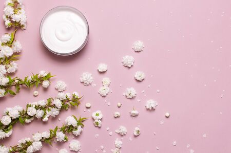 Beauty Spa concept. Opened plastic container with cream and spring White flowers on pink background Flat lay top view. Herbal dermatology cosmetic hygienic cream, organic cosmetic Natural.の写真素材