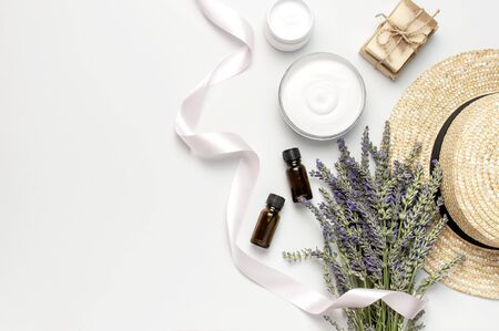 Concept of natural spa cosmetics. Open jar of cream, white cosmetics bottles, soap, straw hat and bouquet of lavender on white background top view copy space. Natural herbal skin care products.の写真素材
