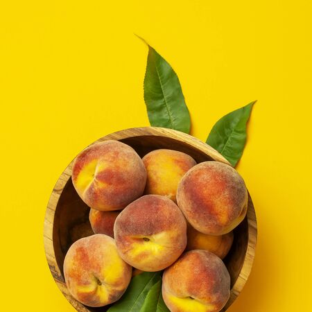 Flat lay composition with peaches. Ripe juicy peaches with green leaves in wooden bowl on yellow background. Flat lay, top view, copy space. Fresh organic fruit, vegan food. Harvest concept.の写真素材