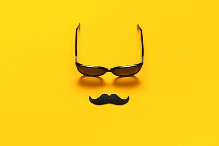 Creative party decoration concept. Black mustache, sunglasses, props for photo booths, carnival parties on yellow background top view flat lay copy space. Father's day Men's health awareness month.の写真素材