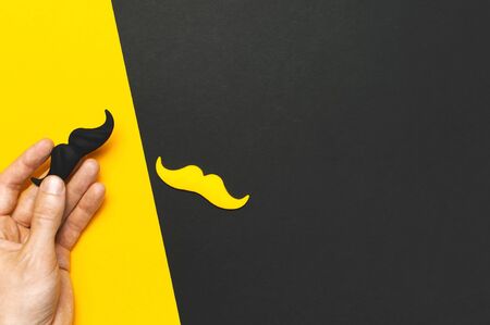 Creative party decoration concept. Male hand holds Black and yellow mustache, props for photo booths, carnival parties on black yellow background top view flat lay Father's day Men's health awareness.の写真素材