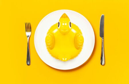 Inflatable mini yellow chicken or duckling on white plate, fork knife on yellow background. Creative food concept, tobacco chicken. Trend Inflatable Children toy for swimming. Layout for design.の写真素材