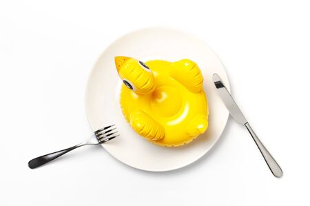 Inflatable mini yellow chicken or duckling on white plate, fork knife on white background. Creative food concept, tobacco chicken. Trend Inflatable Children toy for swimming. Layout for design.の写真素材