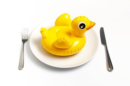 Inflatable mini yellow chicken or duckling on white plate, fork knife on white background. Creative food concept, tobacco chicken. Trend Inflatable Children toy for swimming. Layout for design.の写真素材