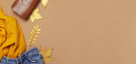 Autumn Fashion Concept. Brown leather women bag, orange knitted sweater, blue jeans, golden autumn leaf on brown background top view flat lay copy space. Fashionable womens accessories Female Clothes.の写真素材