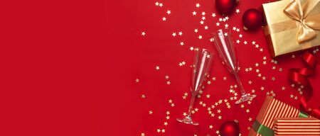 New Year Christmas presents, Christmas balls, champagne glasses, gold confetti stars on red background top view. Flat lay Xmas holiday 2020 celebration. Gift boxes greeting card Festive decorations.の写真素材