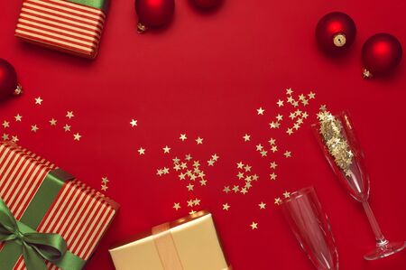 New Year Christmas presents, Christmas balls, champagne glasses, gold confetti stars on red background top view. Flat lay Xmas holiday 2020 celebration. Gift boxes greeting card Festive decorations.の写真素材