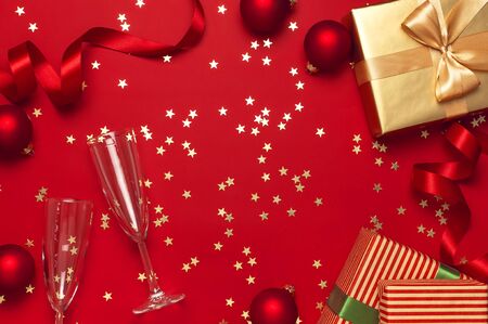 New Year Christmas presents, Christmas balls, champagne glasses, gold confetti stars on red background top view. Flat lay Xmas holiday 2020 celebration. Gift boxes greeting card Festive decorations.の写真素材